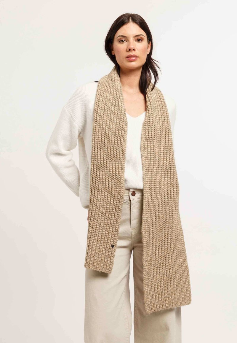 Scarf di maglia beige con una texture a coste, avvolta attorno al collo, abbinata a un maglione bianco e a pantaloni di colore chiaro.