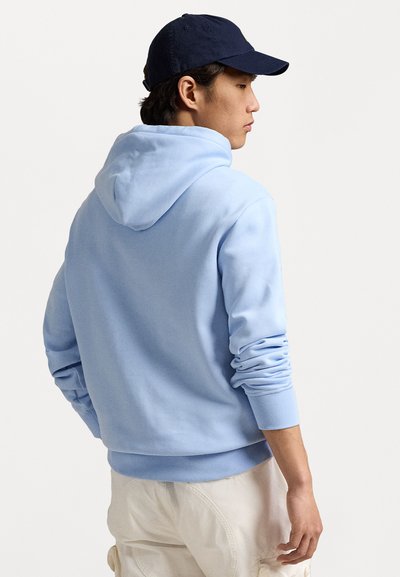 Ανοιχτό μπλε φούτερ με κουκούλα και μαλακή υφή, διαθέτει ριμπ ύφανση στα μανίκια και το στρίφωμα. Φορώντας ένα σκούρο καπέλο, το μοντέλο κοιτάζει μακριά, δείχνοντας την πίσω πλευρά.