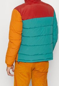 Färgglad pufferjacka med blockfärger i röd, teal och orange, med en quiltad textur och en hög krage för värme.