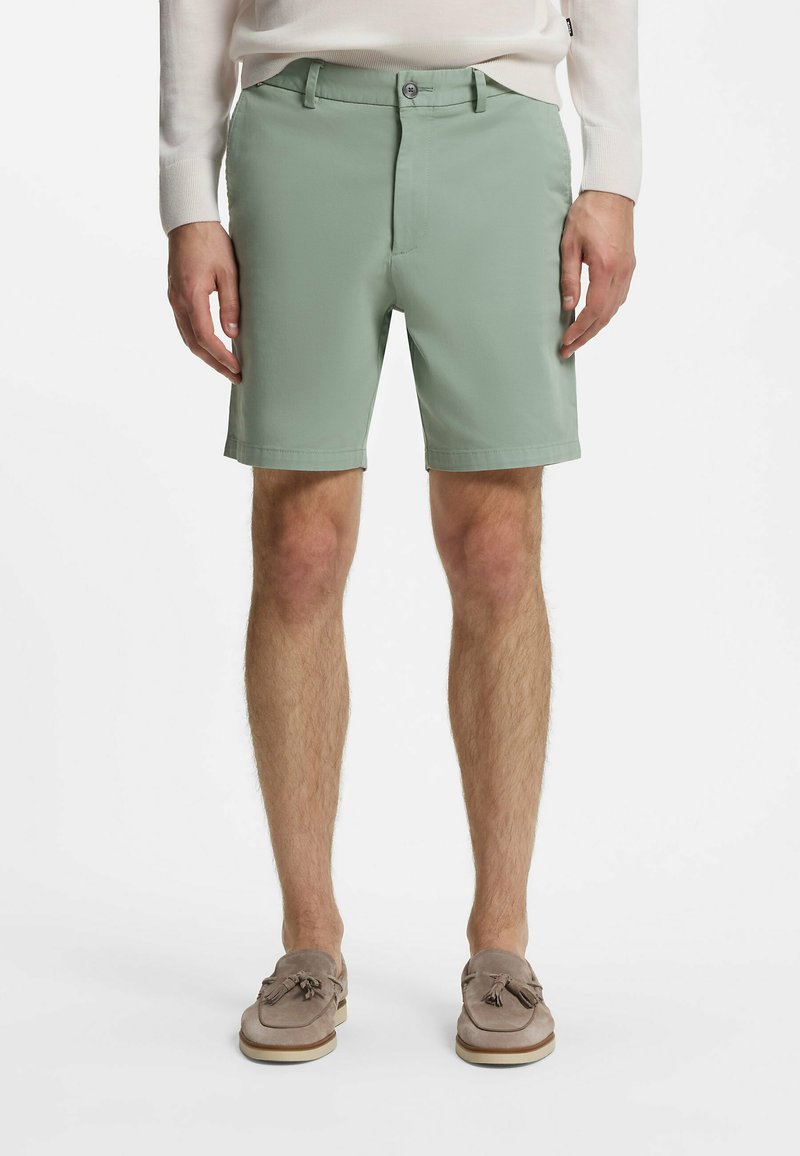 Homme portant un short vert clair sur mesure, des mocassins beige à pampilles et un haut blanc à manches longues, debout devant un fond uni.