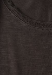 Hnědá látka s hladkým povrchem, která má kulatý výstřih a detail švu ve tvaru cik-cak. Detailní pohled zdůrazňuje jemné tkaní materiálu.