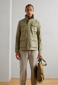 Jachetă utilitară khaki cu patru buzunare frontale, guler înalt și închidere cu buton. Asortată cu pantaloni Bej deschis și purtând o geantă tote bej.