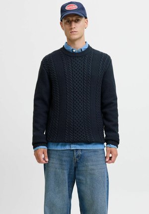 Donkerblauwe gebreide trui met getextureerde kabelpatronen, ronde halslijn en ribgebreide manchetten, gedragen over een chambray shirt, gecombineerd met losse denim.