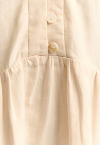 Helles beiges Kleid mit einem plissierten Design und zwei runden Kunststoffknöpfen auf der Vorderseite, aus einem weichen, strukturierten Stoff.