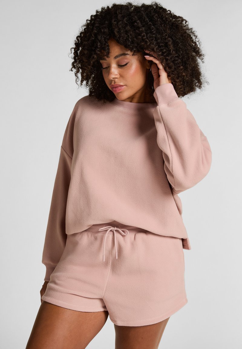 Lichtroze sweatshirt en short set gemaakt van zachte fleece stof. Het sweatshirt heeft een ontspannen pasvorm en licht gezakte schouders, terwijl de shorts zijn voorzien van een verstelbare koord.