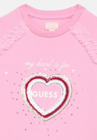Rózsaszín pamut pulóver, középen "GUESS" felirattal díszített, szív alakú flitteres mintával, valamint texturált fodrokkal az vállakon.