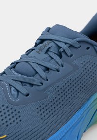 Chaussure de course en maille bleu avec une tige texturée, des œillets renforcés et des lacets détaillés. Elle possède une semelle épaisse et rembourrée aux couleurs dégradées.