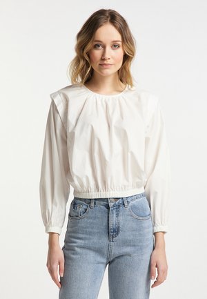 Giovane donna con capelli mossi fino alle spalle che indossa una camicetta bianca a maniche lunghe sbuffate e jeans azzurri a vita alta, in piedi su uno sfondo bianco.