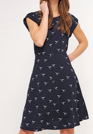 Kobieta w granatowej sukience w białe wzory ptaków, jedną ręką dotyka włosów, a drugą opiera się na biodrze.