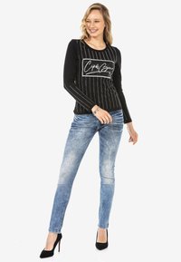 Zwarte lange mouw top met verticale witte strepen en een bedrukte logo; gecombineerd met lichtblauwe skinny jeans en zwarte hakken.