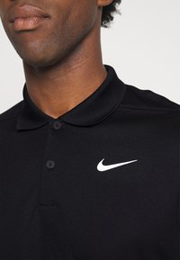 Crna polo majica s ovratnikom, dva gumba i bijelim Nike swoosh logotipom na lijevoj strani prsnog koša. Izrađena od teksturirane tkanine.