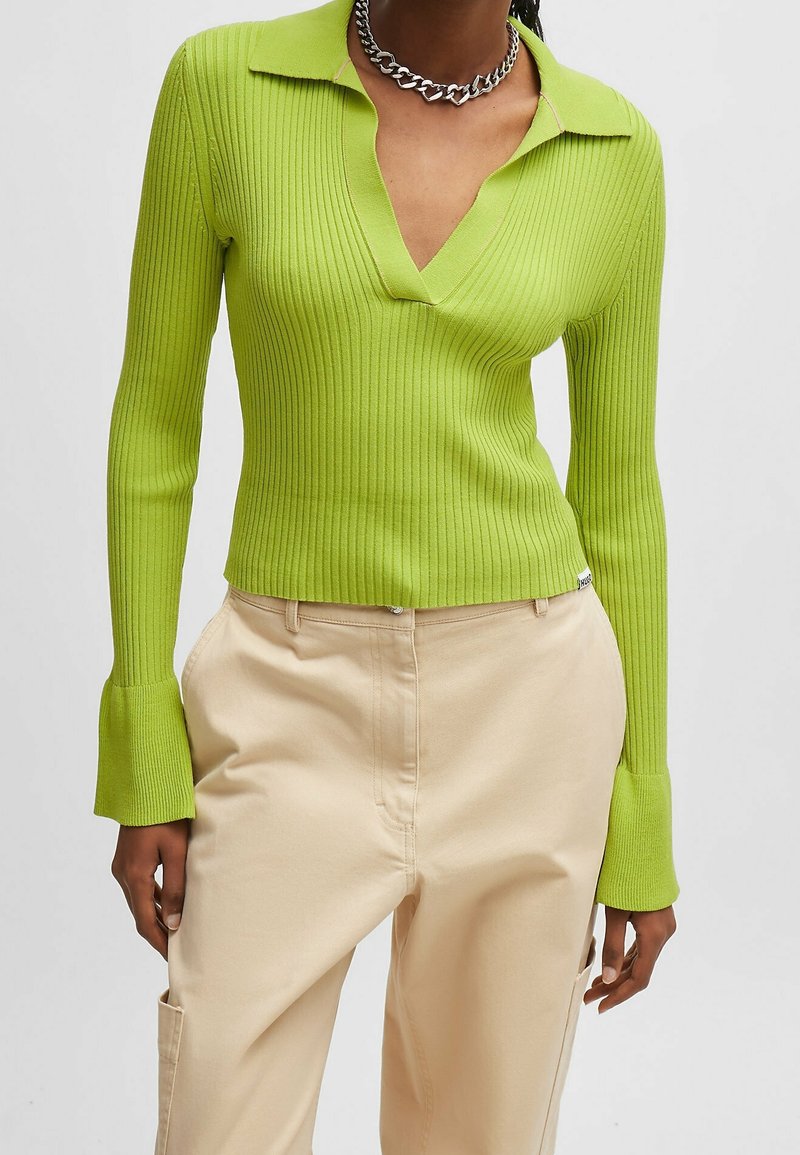 Zielony prążkowany top z długim rękawem, z kołnierzykiem w serek i rozkloszowanymi mankietami, w połączeniu z beżowymi spodniami. Posiada dopasowany krój i teksturowany materiał.