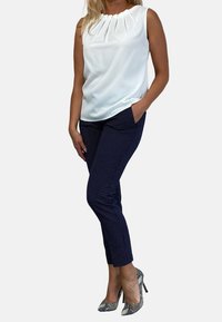 Kvinna som står med händerna i fickorna iförd en vit ärmlös blus, marinblå slim fit-byxor med små prickar, och högklackade skor med ormskinnsmönster.