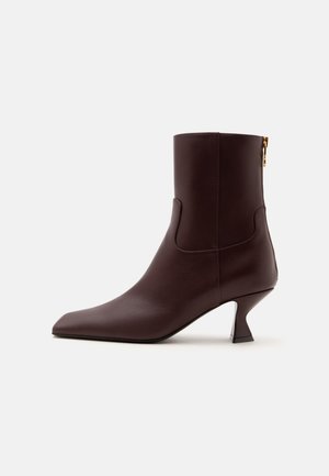 Bottine en cuir brun foncé avec bout pointu, talon kitten incurvé et fermeture éclair à l'arrière sur un fond clair uni.