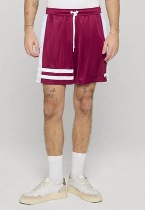 Mann trägt bordeauxrote Sportshorts mit weißen Seitenstreifen und weißen Turnschuhen, steht vor einem einfarbigen Hintergrund.