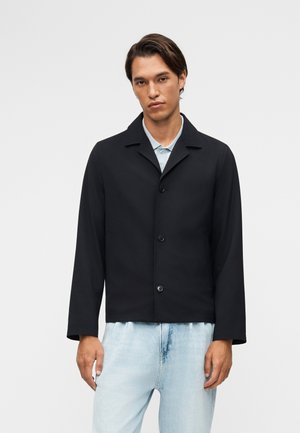 Mand iført en sort knapjakke over en lyseblå skjorte med krave og lyseblå jeans, stående foran en ensfarvet lys baggrund.