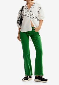 Camisa blanca con texto negro, pantalones de mezclilla verdes acampanados y zapatillas negras. La camisa tiene un ajuste holgado y una silueta oversized.
