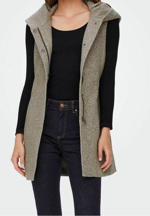 Długa, szarozielona kamizelka bez rękawów z kapturem, posiadająca z przodu zatrzaski i dwie boczne kieszenie, noszona na czarnej bluzce i ciemnych dżinsach.