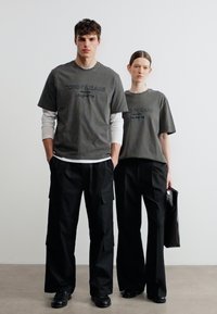 Νεαρός άνδρας και γυναίκα στέκονται δίπλα δίπλα, φορώντας σκουρόχρωμα υπερμεγέθη T-shirts, μαύρο φαρδύ παντελόνι και μαύρα παπούτσια· η γυναίκα κρατά μαύρη τσάντα.