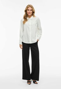 Camicetta bianca con colletto, abbinata a pantaloni neri a gamba larga. L'outfit presenta una vestibilità comoda e una texture liscia. Includerà sandali.