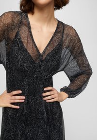 Robe noire transparente avec un motif paisley, décolleté en V profond, manches ballon longues et fermeture à boutons sur le devant, fabriquée dans un tissu doux et texturé.
