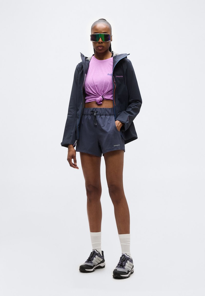 Femme portant des lunettes de soleil miroir oversize, une chemise violette nouée, une veste marine, un short marine, des chaussettes blanches et des chaussures de sport grises, debout devant un fond blanc.