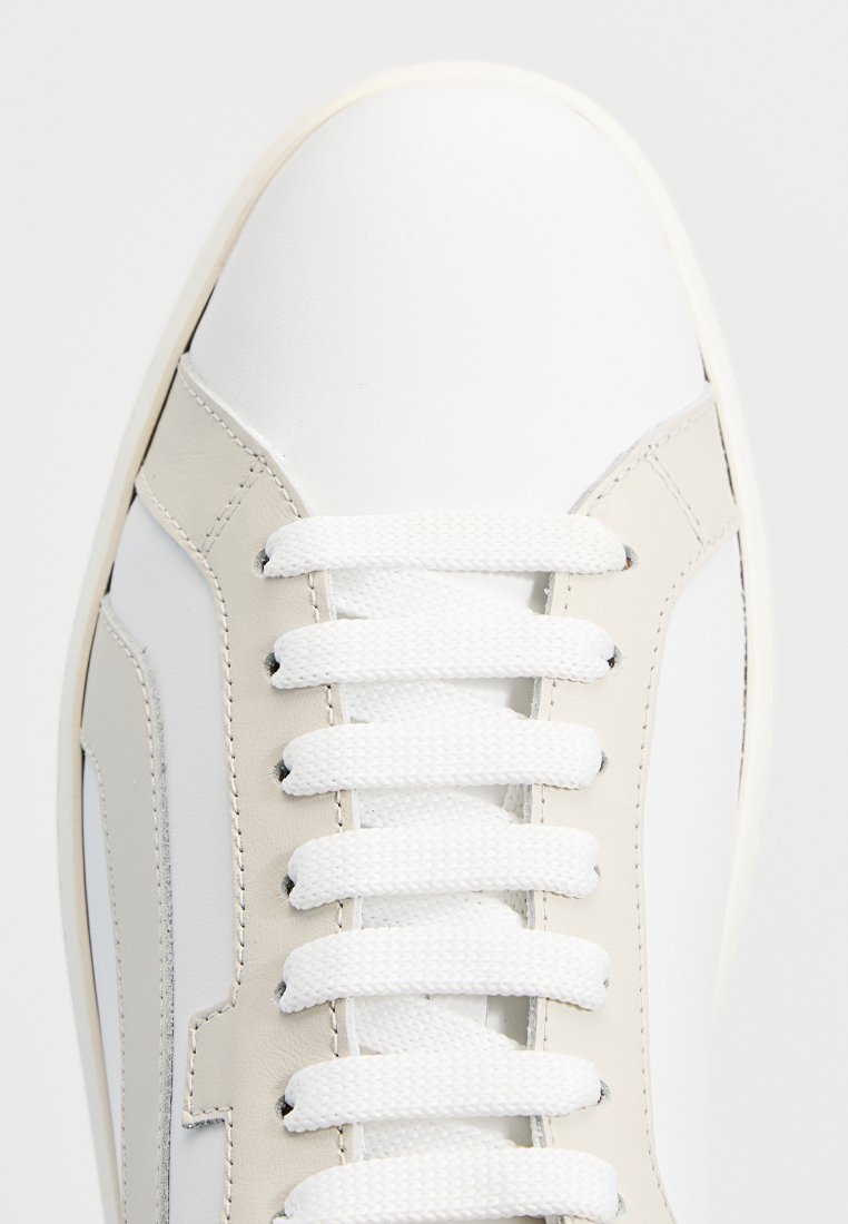 Białe skórzane sneakersy z beżowymi akcentami, płaskimi białymi sznurowadłami i zaokrąglonym czubkiem. Charakteryzują się gładką teksturą i solidną podeszwą.