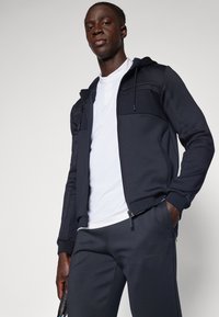 Mørkeblå hettegenser med glidelås og teksturerte skulderpaneler, kombinert med matchende mørke bukser. Antrekket inkluderer en hvit undertrøye.