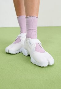 White sneakers with textured material, elastic straps, and distinctive toe design, paired with light pink ribbed socks. Green floor background.