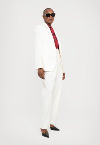 Witte op maat gemaakte pak met een rood en zwart gestreepte das. Zwarte zonnebril en spitse zwarte hakken maken de look compleet. Glad textuur en gestructureerd ontwerp.