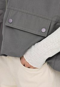 Harmaa liivi etutaskuilla, jossa on violetit napit ja kontrastiväriset ompeleet. Valkoinen teksturoitu pitkähiainen paita alla, yhdistettynä kermanvärisiin housuihin.