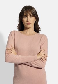 Světle růžový svetr s dlouhými rukávy a žebrovanými manžetami. Měkká textura, zakulacený výstřih a volný střih. Modelka pózuje se zkříženýma rukama.