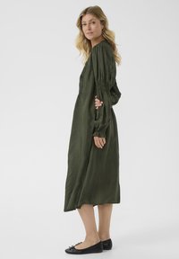 Abito lungo color verde oliva con spalle arricciate e maniche lunghe. Realizzato in un tessuto leggero, presenta un orlo lungo fino al pavimento e un design semplice.