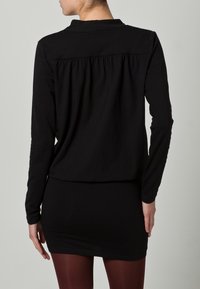 Svart långärmad topp med krage, rynkad baksida och slät yta, kombinerad med en figursydd svart kjol och vinröda strumpbyxor.