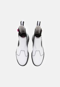 Bílé kožené kotníkové boty s černými gumovými podrážkami, s elastickými bočními částmi a poutky v kontrastních barvách. Podrobnosti o zesíleném šití.