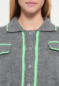 Hall halli villane kardigan roheliste detailidega, varustatud krae, kahe eesmise tasku ja hallide nuppudega keskel. Tekstuuriga kootud kangas.