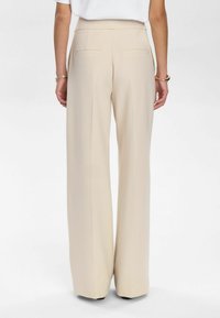 Pantaloni beige a gamba larga con una vestibilità sartoriale, dotati di due tasche posteriori e una texture di tessuto liscia, che terminano sopra le calzature.
