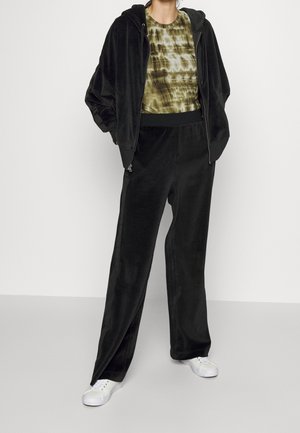 Personne portant une veste à capuche zippée en velours noir et un pantalon, avec un t-shirt tie-dye vert et des baskets blanches, debout devant un fond uni.