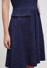 Femme portant une robe bleu foncé avec un motif floral damassé subtil et un détail peplum à la taille, debout devant un fond uni.
