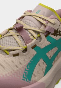 Nærbillede af beige og pink ASICS-sneaker med snørebånd med gule accenter og turkis side-logo, der viser mesh-øvre og polstret tunge.