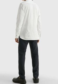 Camicia bianca a maniche lunghe con orlo dritto, indossata con pantaloni scuri a fantasia e scarpe nere. Presenta polsini con bottoni e una vestibilità comoda.
