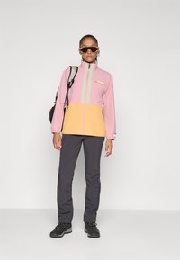 Mujer con chaqueta rosa y amarilla, pantalones gris oscuro, botas de senderismo negras, gafas de sol y llevando una mochila, de pie frente a un fondo liso.