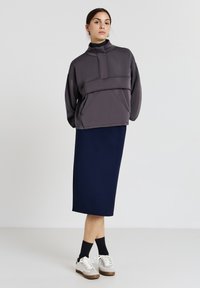 Pullover grigio oversize con collo alto e tasca frontale, abbinato a una gonna blu navy lunga fino al ginocchio. Il modello indossa sneaker grigie e bianche.