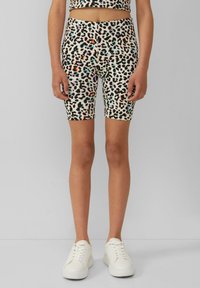 Vrouw in een bijpassende leopardprint broek en top met zwarte, oranje, lila en blauwe vlekken, gecombineerd met witte sneakers op een grijze achtergrond.