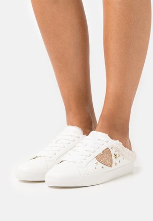 Vita slip-on sneakers med guld hjärtan och stjärnspik, fluffig innerfoder, som bärs på bara fötter framför en enfärgad bakgrund.