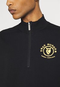 Černá zipová mikina s vysokým límcem a žlutým kruhovým logem "Tree Hugger" s designem ptáčka. Hladký povrch látky.