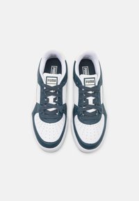 Para białych i ciemnoniebieskich trampek Puma z sznurowadłami, perforowanym czubkiem i logo na języku, widziana z góry na białym tle.