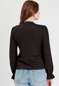 Blouse noire à côtes avec de longues manches bouffantes, présentant des poignets froncés et un col rond. Le tissu a une texture douce, portée avec un jean clair.