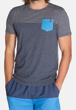 Mężczyzna ubrany w szarą koszulkę z krótkim rękawem o wzorze, z jasnoniebieską kieszenią na piersi i dwukolorowe niebieskie szorty, z rękami w kieszeniach.