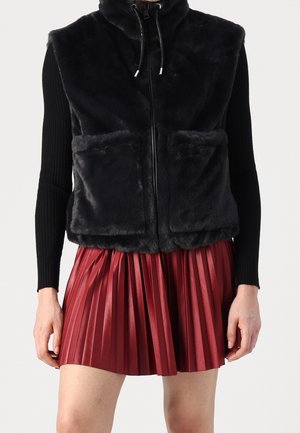 Veste sans manches en fausse fourrure noire zippée avec poches avant, portée sur des manches longues noires et une mini-jupe plissée rouge.